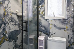 A bathroom with a glass shower enclosure, a white sink, and a marble-patterned wall. - Studio Sleeps 2 With Parking (Thornton Heath)