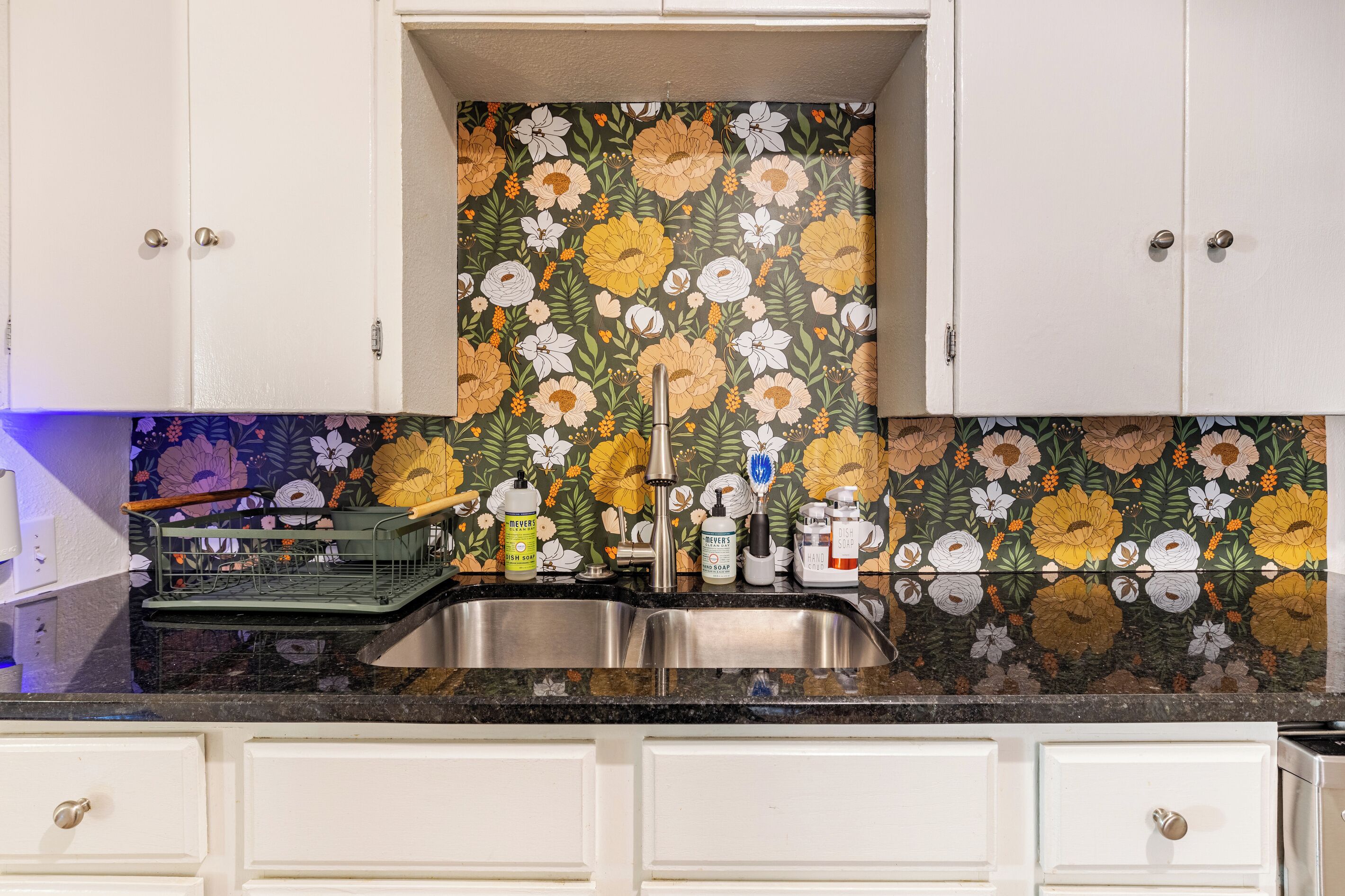 Una cocina con salpicadero floral, pileta de acero inoxidable y armarios blancos.