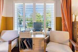 Ein Zimmer mit Schreibtisch, zwei Stühlen, einem Fernseher und einem Fenster mit Jalousien.