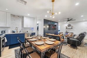 A modern kitchen with blue cabinets, a dining area with a wooden table set for four, and a living room with a sofa and a TV.