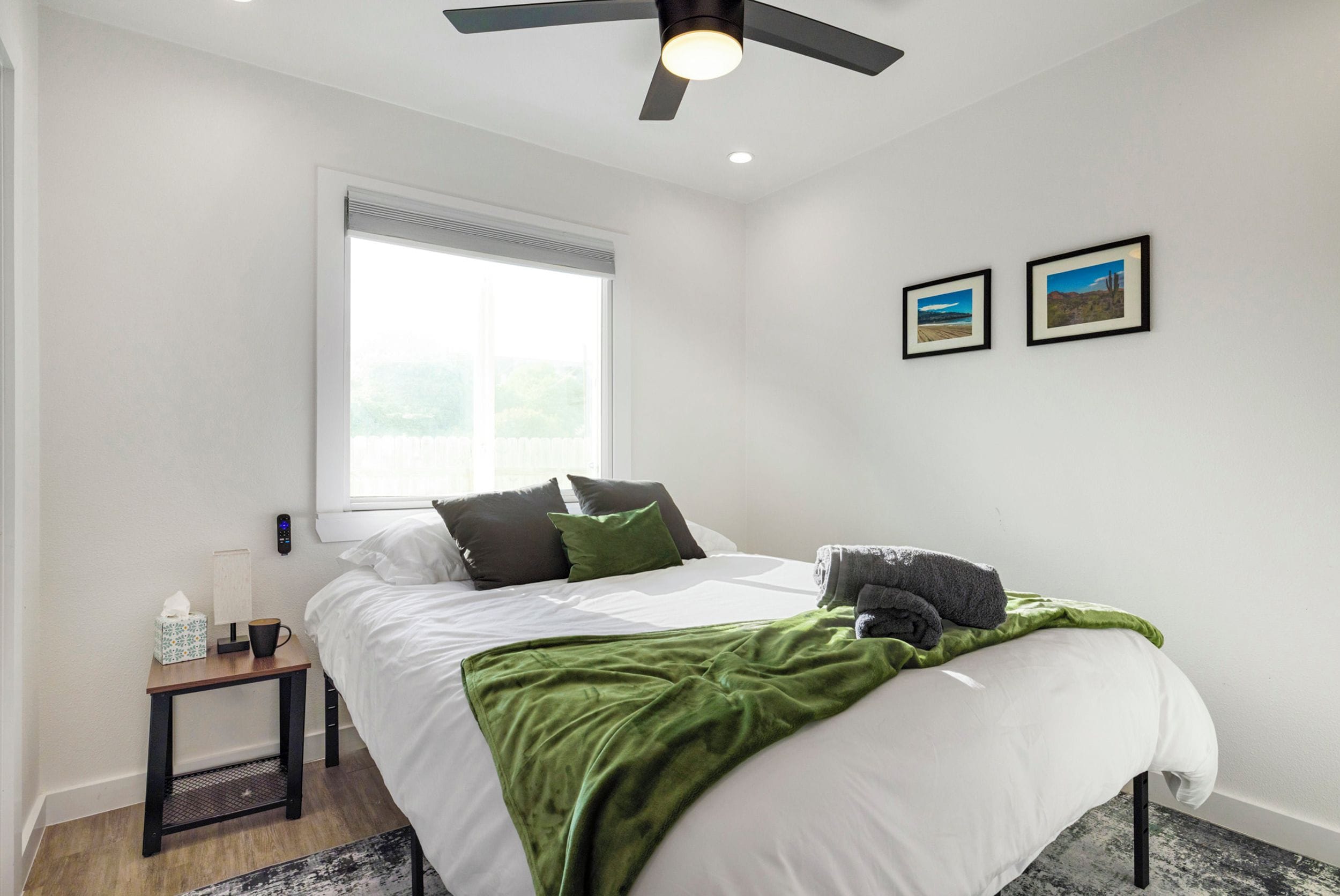 A bedroom with a bed, a bedside table, a nightstand, a ceiling fan, a window, and two framed pictures on the wall.