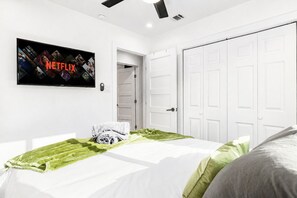 A bedroom with a bed, a television displaying the Netflix logo, white walls, and a white door.