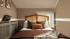A bedroom with a bed, pillows, a bedside table with a lamp, a plant, and a window with blinds. - Charming Countryside Cottage (Blandford Forum)