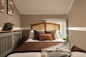 Ein Schlafzimmer mit einem Bett, Kissen, einem Nachttisch mit Lampe, einer Pflanze und einem Fenster mit Jalousien.