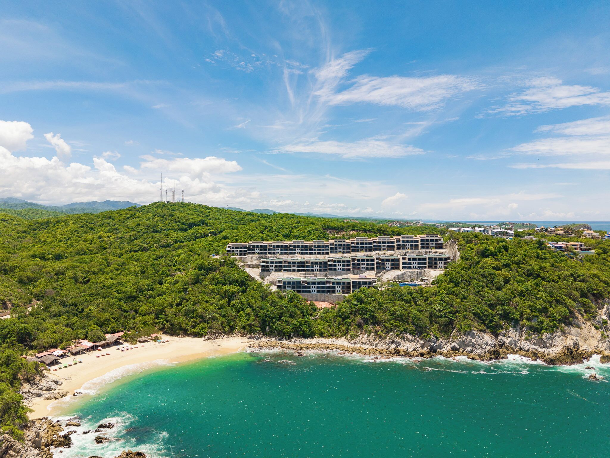 A coastal hotel with a large building surrounded by dense greenery, a beach, and clear turquoise waters.