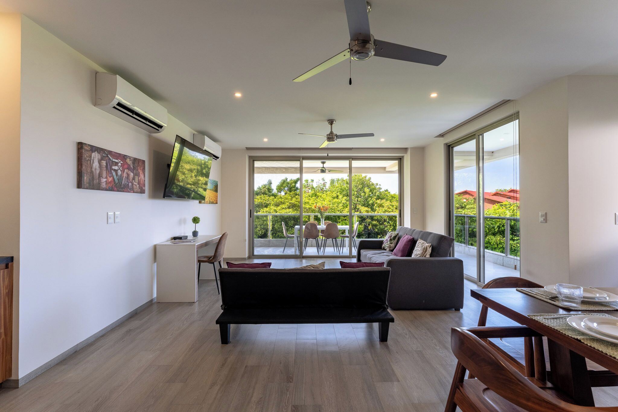 A spacious living area with a sofa, dining table, and a balcony with a view of greenery.