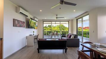 A spacious living area with a sofa, dining table, and a balcony with a view of greenery.
