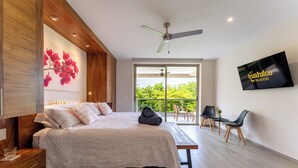 A modern bedroom with a bed, a desk, a chair, a TV, and a balcony with a view of greenery. - 2nd Level 2BR | Oceanview & Beach Access (Huatulco)