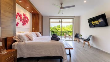 A modern bedroom with a bed, a desk, a chair, a TV, and a balcony with a view of greenery.