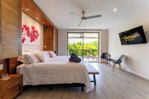 Ein modernes Schlafzimmer mit Bett, Schreibtisch, Sessel, Fernseher und einem Balkon mit Blick auf Grünflächen.
