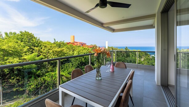 A balcony with a table, chairs, a vase with flowers, and a view of the ocean and greenery. - 2nd Level 2BR | Oceanview & Beach Access (Huatulco)