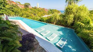 A swimming pool with lounge chairs, surrounded by lush greenery and overlooking a coastal area.