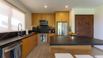 A modern kitchen with wooden cabinets, a stainless steel refrigerator, a microwave, and a large island with a bowl of fruit.