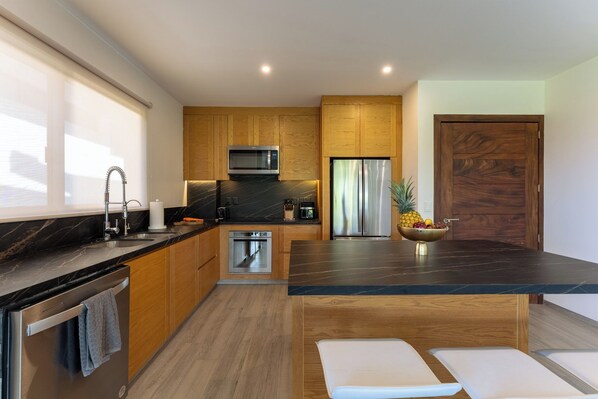 A modern kitchen with wooden cabinets, a stainless steel refrigerator, a microwave, and a large island with a bowl of fruit.