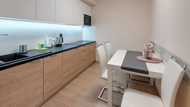 A modern kitchen with a black countertop, wooden cabinets, a white dining table with two chairs, and a small vase with flowers on the table. - RentPlanet - Apartament Triventi (Karpacz)