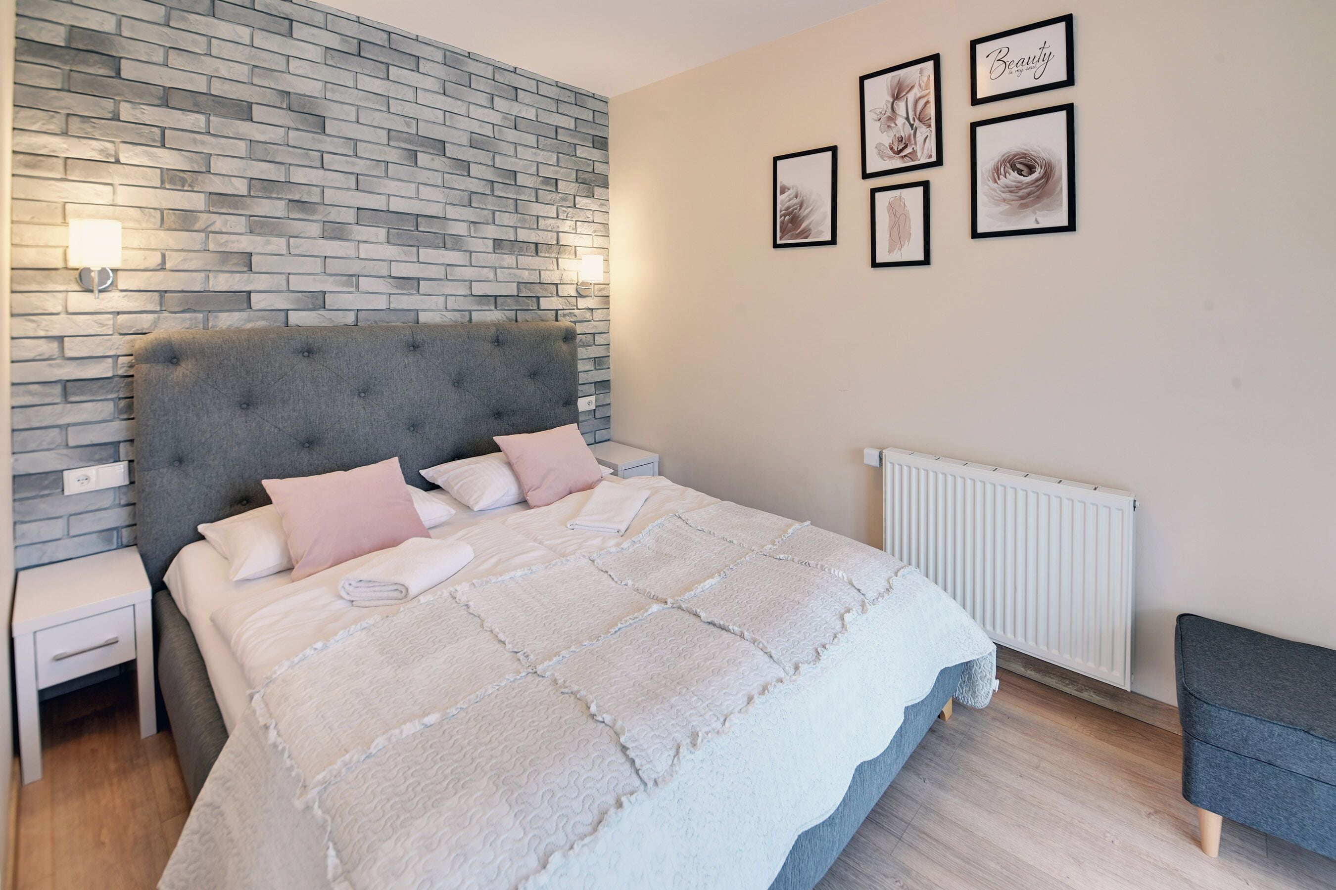 Ein Schlafzimmer mit einem Kopfende aus Ziegelsteinoptik, einem Bett mit weißer Bettdecke, einem Nachttisch, einer Wandleuchte und gerahmten Kunstwerken.