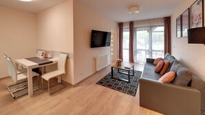 A modern living room with a sofa, a dining table with chairs, a flat-screen TV mounted on the wall, and a large window with curtains. - RentPlanet - Apartament Triventi (Karpacz)