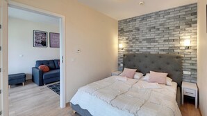 Un dormitorio con una cama grande, cabecera gris, pared de piedra y una pequeña zona de estar con sofá y cojines.