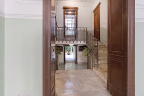 Un pasillo con suelo de mármol que conduce a una escalera con barandilla de hierro forjado y una puerta de madera a la derecha.