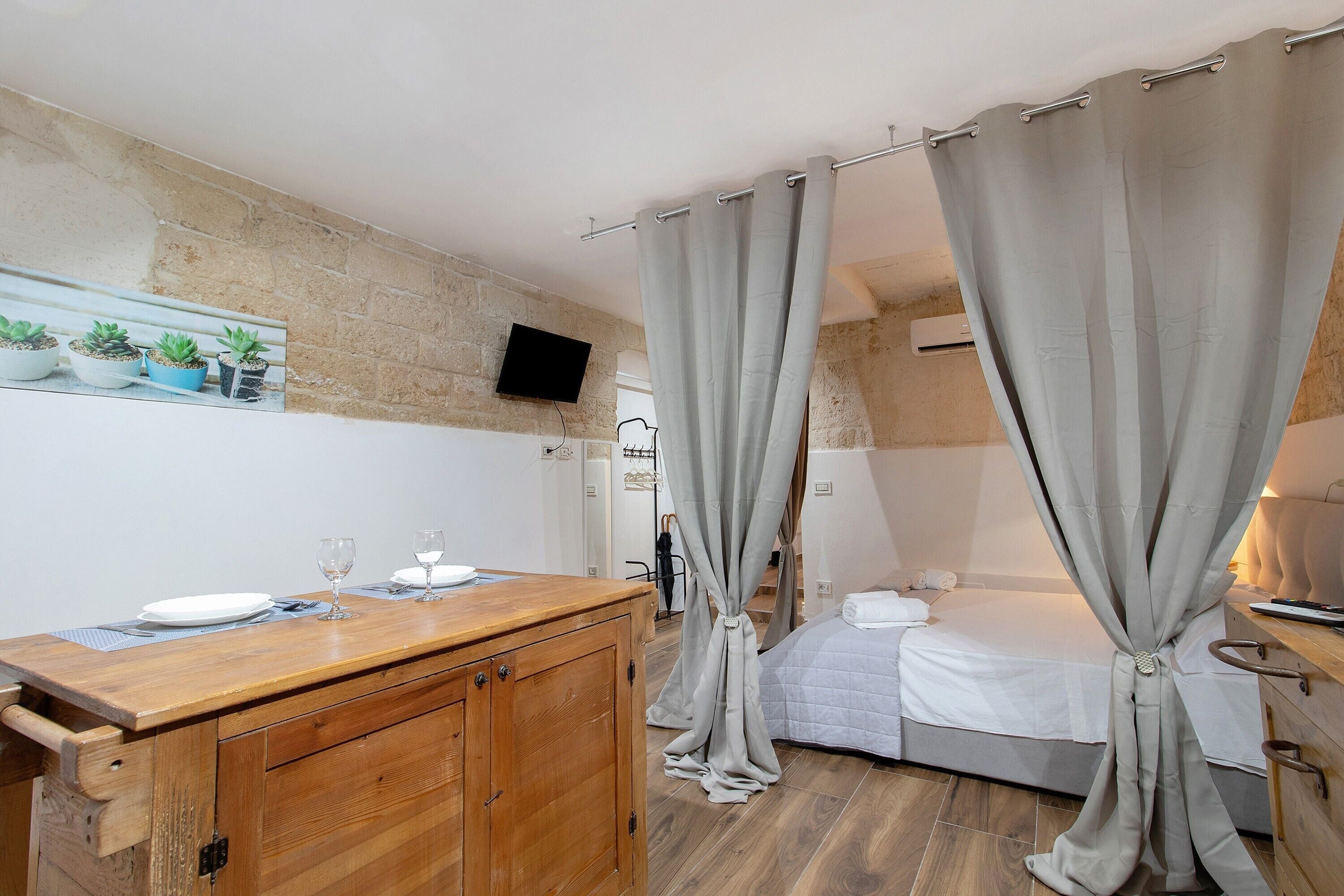 A bedroom with a bed, a wooden dresser, a television mounted on a brick wall, and a window with curtains.