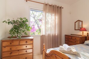 A bedroom with a wooden dresser, a bed with a rolled towel, a bedside lamp, and a window with curtains. - Villa Nadezda – 3-Bedroom Villa with Pool near Paphos Harbour (Pafos)