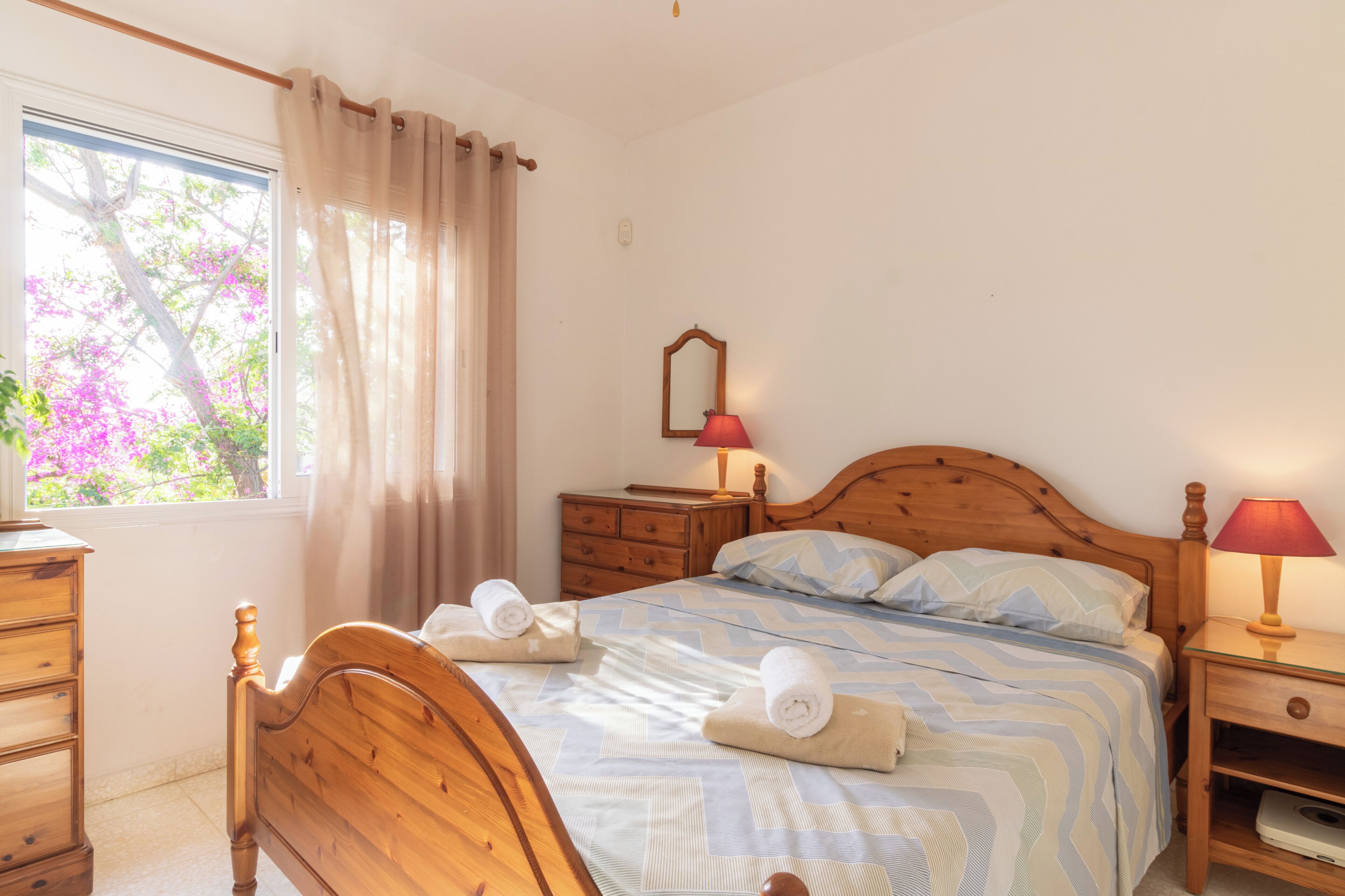 A bedroom with a wooden bed, two bedside tables with lamps, a dresser, and a window with curtains.