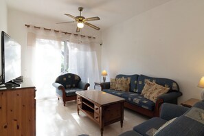 Ein Wohnzimmer mit einer Couch, Sesseln, einem Couchtisch, einem Fernseher und einem Deckenventilator.