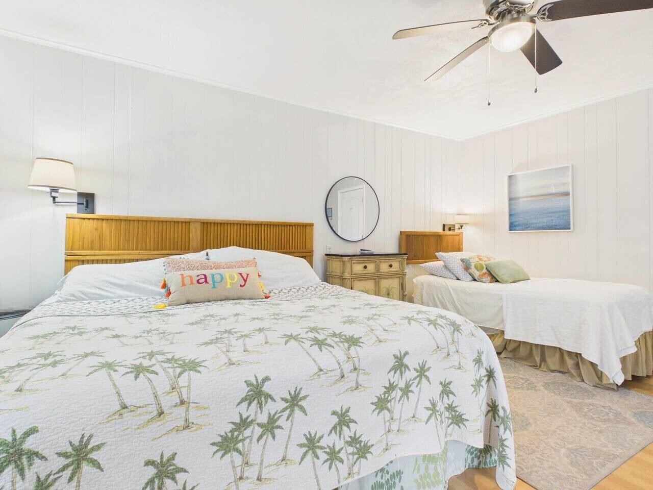 A bedroom with a large bed, two smaller beds, a ceiling fan, a round mirror, and a painting on the wall.