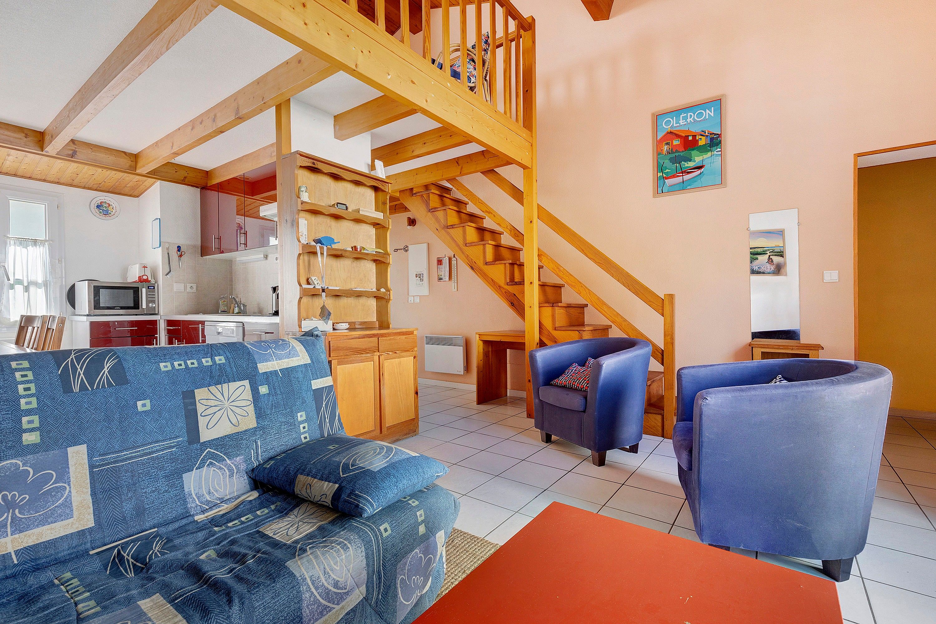 Un salon avec un escalier en bois, un canapé, deux fauteuils et, au fond, un coin cuisine.