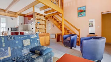 Sala de estar com escada de madeira, um sofá, duas poltronas e uma área de cozinha ao fundo.