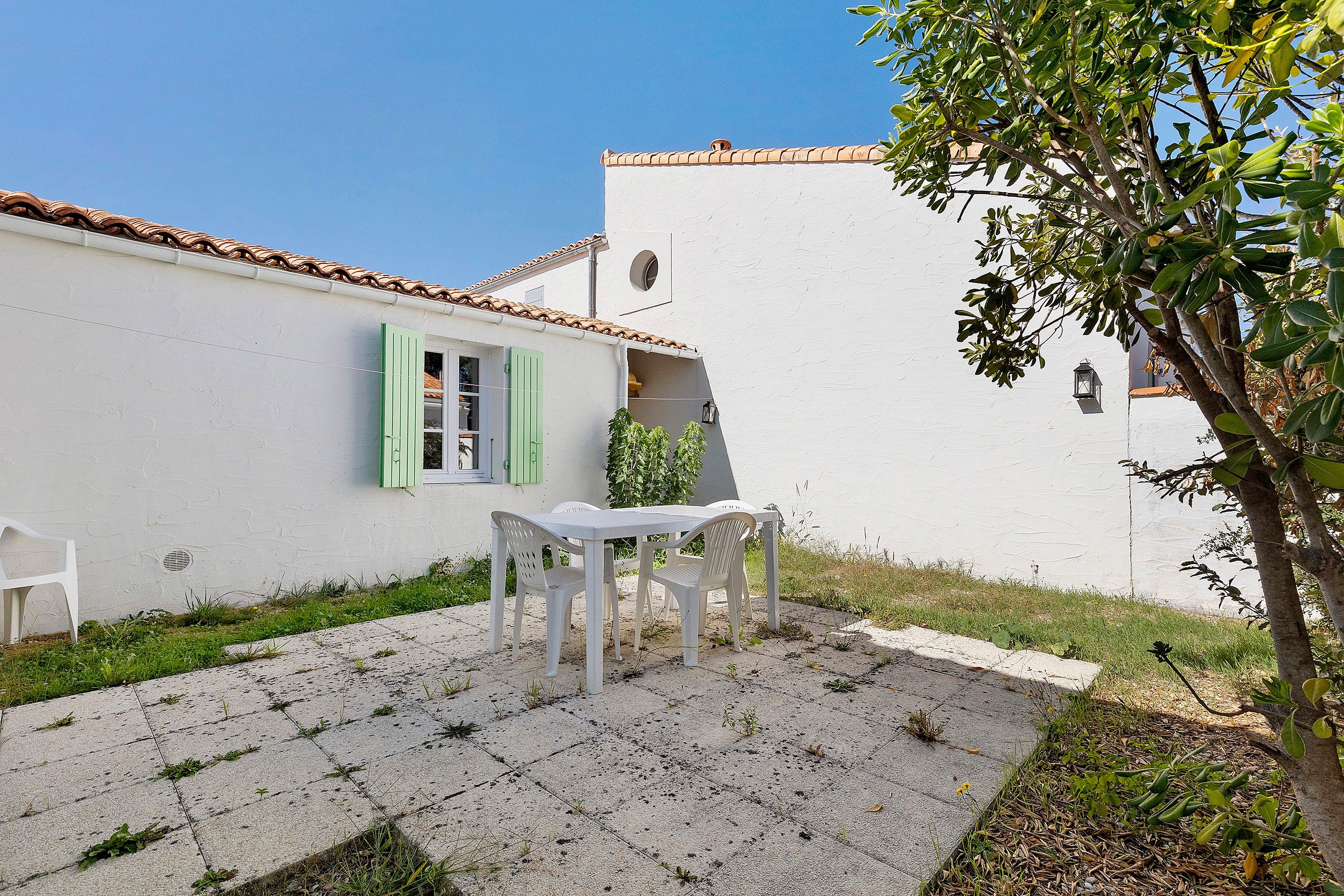 Un bâtiment blanc avec un toit recouvert de tuiles, une petite table avec des chaises et un arbre au premier plan.