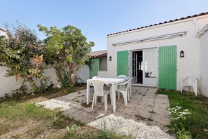 Uma área de pátio com mesa e cadeiras, um edifício branco com persianas verdes e uma árvore com folhas verdes.