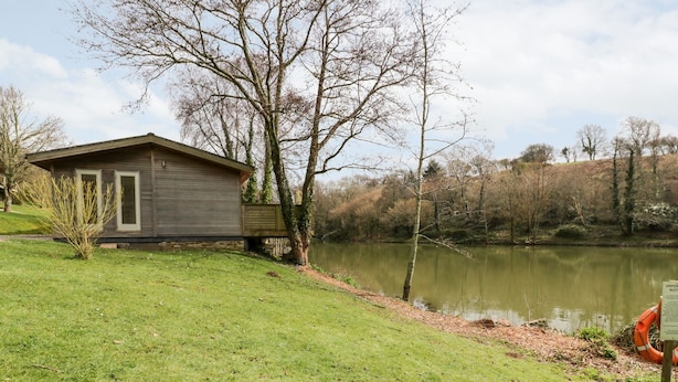 Una cabaña de madera junto a un lago, con un área verde al frente y árboles en el fondo.