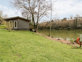Una cabaña de madera junto a un lago, con un área verde al frente y árboles en el fondo.