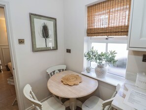 Ein kleiner Essbereich mit einem runden Tisch, weißen Stühlen, einem gerahmten Bild an der Wand, einem Fenster mit Aussicht und einem Blumentopf auf der Fensterbank.