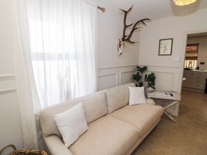 Un canapé beige avec des coussins blancs, dans une pièce dotée d’une grande fenêtre, d’une tête de cerf accrochée au mur et d’une petite table sur laquelle est posée une plante.