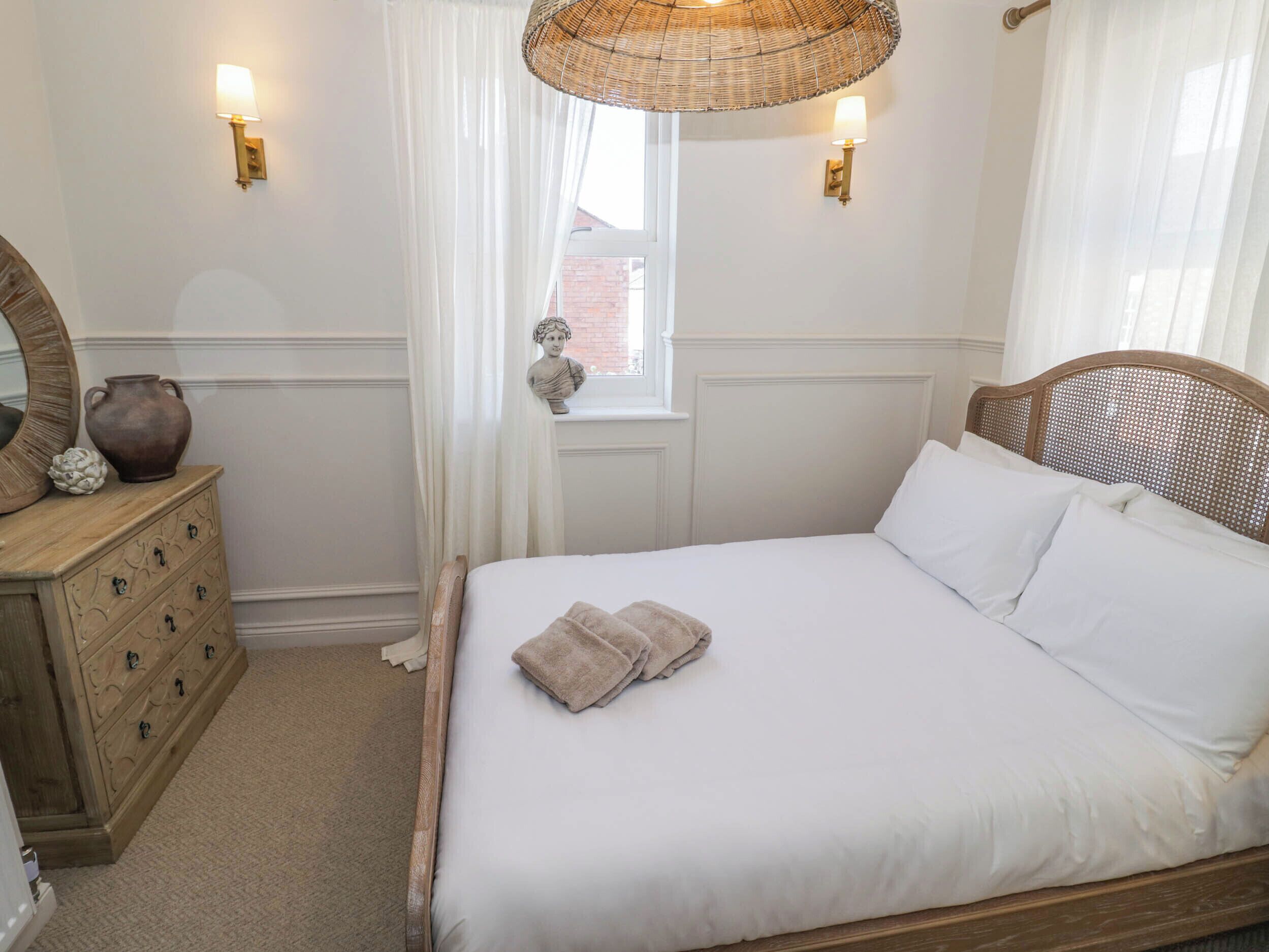 A bedroom with a wooden bed, white bedding, a dresser with drawers, a mirror, a vase, and two wall-mounted lamps.