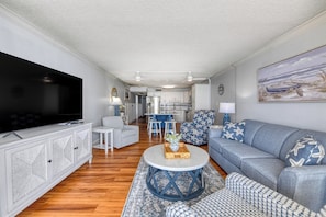 A living room with a television, sofa, and chairs. There is a kitchen in the background. - Royal Garden 1115 - 2 Bed 2 Bath Luxury Condo with Ocean and Marsh Views! (Garden City Beach)