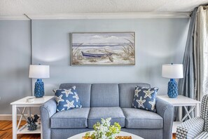 Ein Wohnzimmer mit grauem Sofa, Kissen mit Sternenmuster, einem Bild von einem Boot am Strand und zwei Lampen auf Nachttischen.