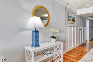 A hallway with a white console table, a blue lamp, a mirror, a framed picture, and a potted orchid. - Royal Garden 1115 - 2 Bed 2 Bath Luxury Condo with Ocean and Marsh Views! (Garden City Beach)