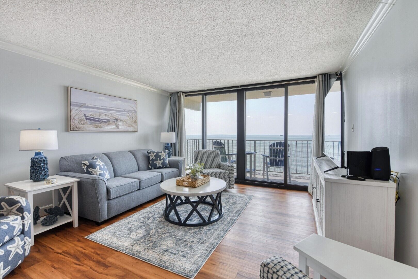 Ein Wohnzimmer mit einer Couch, einem Couchtisch und Sesseln, ein Balkon mit Meerblick und ein Fernseher auf einem Schrank.