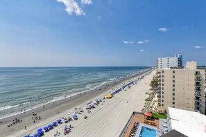 A beach with a long stretch of umbrellas and lounge chairs, a clear sky, and a building with balconies overlooking the beach. - Royal Garden 1115 - 2 Bed 2 Bath Luxury Condo with Ocean and Marsh Views! (Garden City Beach)