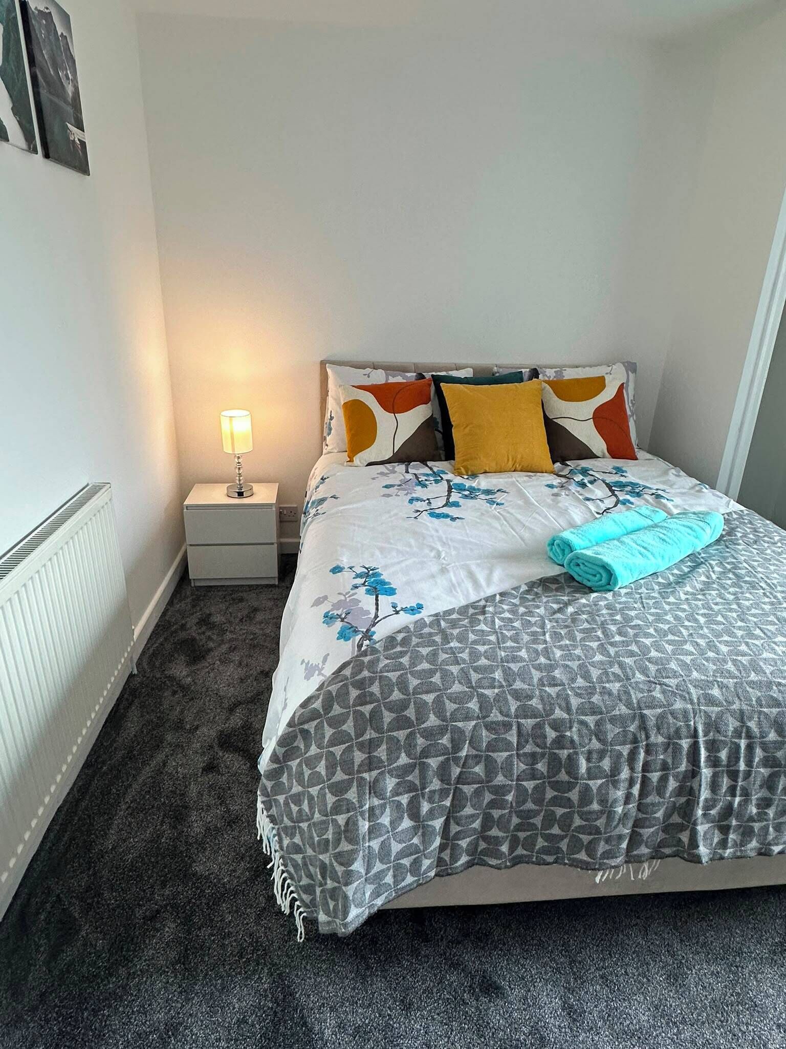 A neatly made bed with a floral bedspread, a rolled towel, and a bedside lamp on a nightstand.