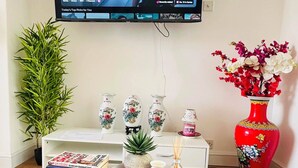 Ein Wohnzimmer mit Fernseher, der eine Serie zeigt, einem weißen Couchtisch mit Büchern und Fernbedienung, einer roten Vase mit Blumen und einer Topfpflanze.