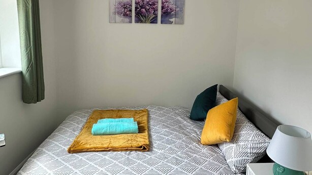 Ein Schlafzimmer mit Bett, Nachttisch, Lampe, einem Fenster mit Vorhängen und Wandbildern.
