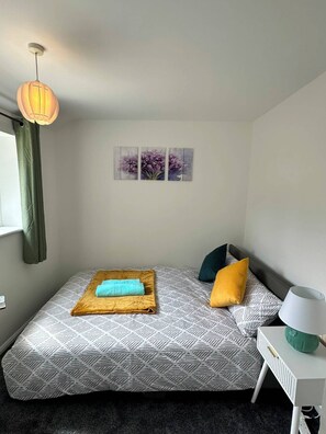 A bedroom with a bed, a bedside table, a lamp, a window with curtains, and wall art. - Everest Cozy Corner (Colchester)