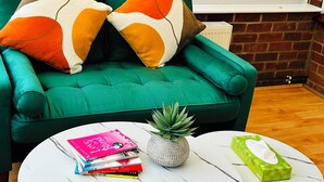 Ein grünes Sofa mit zwei Zierkissen, ein kleiner runder Tisch mit einem Buch und einer Pflanze sowie eine grüne Papiertauschbox.