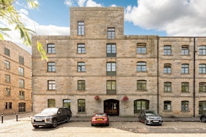 A multi-story stone building with a central entrance, several windows, and a parking area with cars. - Historic Whisky Bond 2BR | Free Parking - Pass the Keys (Edinburgh)