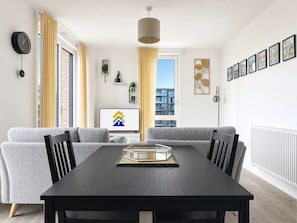 Ein modernes Wohnzimmer mit einem grauen Sofa, einem schwarzen Esstisch und einem großen Fenster mit Vorhängen.
