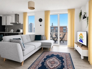 Ein modernes Wohnzimmer mit einem grauen Ecksofa, einem gemusterten Teppich, einem Fernseher und einem Balkon mit Sitzgelegenheit.